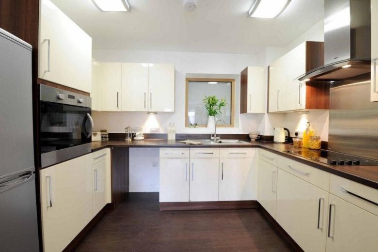 A bright, modern kitchen with a U-shaped design. The cabinets are a combination of cream and dark brown wood, with a white countertop and a stainless steel sink. A framed mirror hangs on the wall, and a potted plant sits on the counter. Appliances include a refrigerator, oven, and extractor fan.