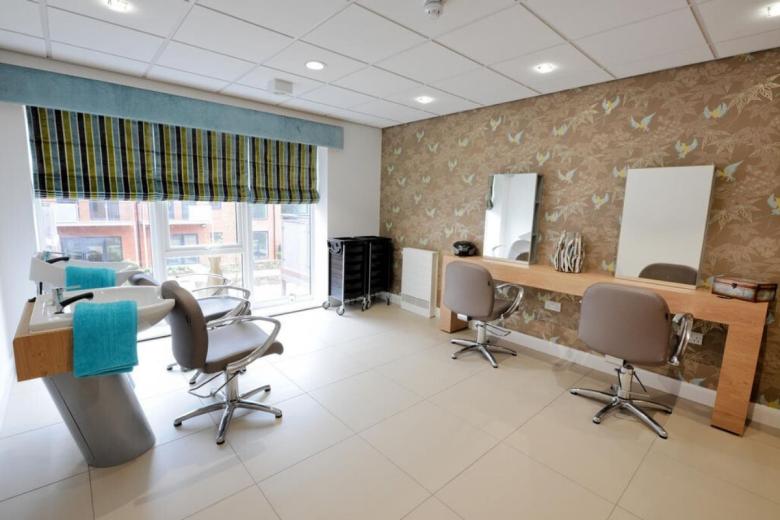 Hair salon with three chairs, a washbasin, and a mirrored vanity. A black trolley sits to the left, and a striped blind is positioned above the window. The wall is covered in a wallpaper featuring brown and beige birds.
