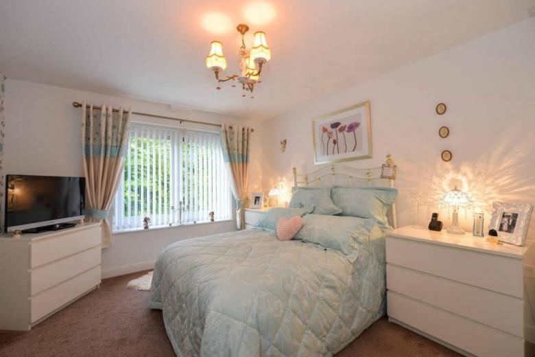 A well-appointed bedroom featuring a light-colored bed with a pale blue quilted bedspread and pillows. A decorative floral print artwork hangs above the bed. A television sits on a white dresser to the left, while a nightstand with a lamp and framed photograph is positioned on the right. Curtains with a floral pattern are drawn, and a chandelier provides ambient lighting. The room has a warm, inviting atmosphere.