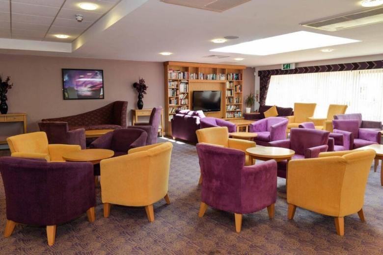 A brightly lit lounge area featuring a collection of purple and yellow armchairs arranged around small tables. A large bookcase filled with books is positioned against one wall, and a television is mounted on the wall opposite. The room has a neutral-toned carpet and a patterned window blind.
