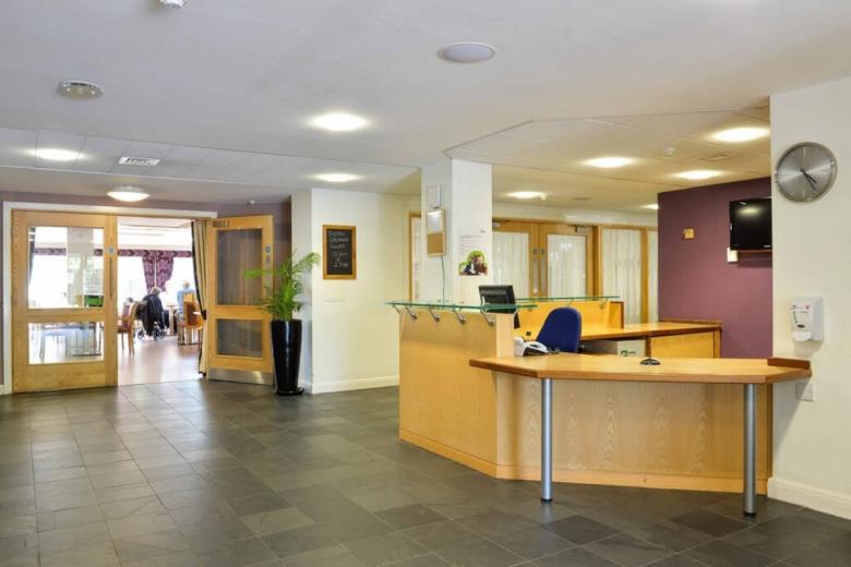 A reception area with a curved wooden desk and computer. Behind the desk, a black plant sits on a dark pot. A purple wall is visible in the background, along with a television and a wall-mounted clock. Through a glass door, a room filled with seated individuals is visible, and a black chalkboard with a picture is present on the wall to the left.