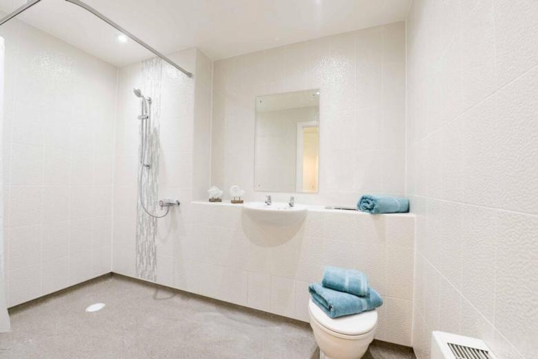 A modern, accessible bathroom features a walk-in shower with a linear showerhead and a partially tiled wall. A white sink with a matching vanity and a toilet are positioned adjacent to the shower. A large, rectangular mirror hangs above the vanity. Three folded blue towels are stacked on the toilet seat. The walls and floor are covered in white tiles.