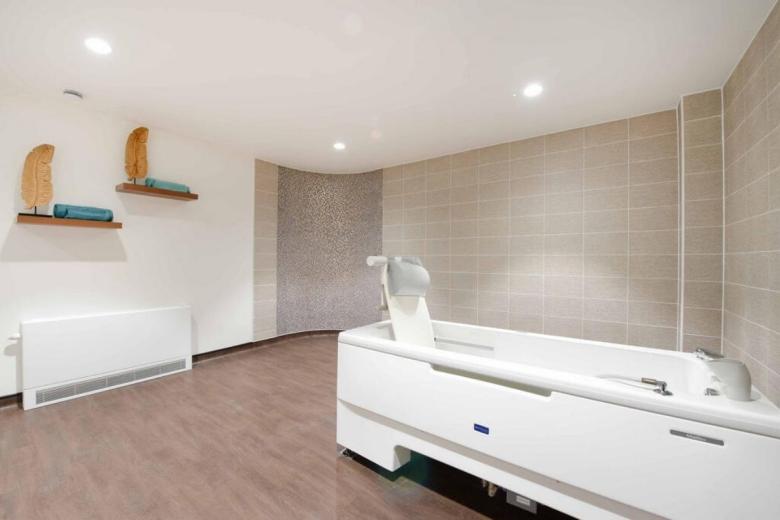 A modern, spacious bathroom features a white, reclining bathtub positioned centrally in a room with light brown tiled walls and a textured wall paneling. Two blue towels are draped on floating wooden shelves to the left. A white radiator is located on the right side of the frame. The lighting is bright and even, illuminating the room.