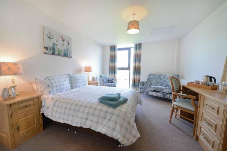 A well-appointed bedroom featuring a double bed with a light blue and white checkered duvet. A bedside table with a lamp and a small decorative item sits on one side of the bed, while a small armchair and a desk with a lamp and a small table with a glass of water are visible in the background. The walls are painted a neutral white, and a window with curtains is located on the right side of the room.