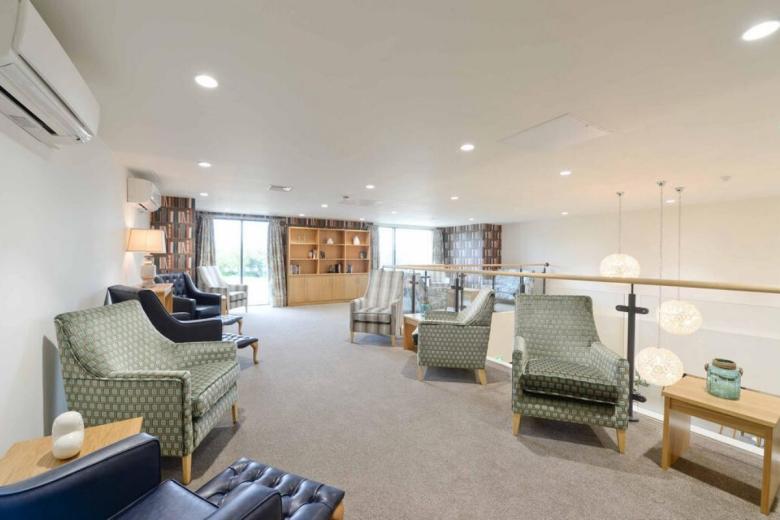 A bright, spacious lounge area with a light grey carpet. Several upholstered armchairs with a green and white patterned fabric are arranged around the room. A wooden coffee table with a candle sits in front of one chair. A low, decorative railing separates the lounge from a mezzanine level above, which features a bookshelf. A white air conditioning unit is mounted on the wall, and several pendant lights hang from the ceiling.