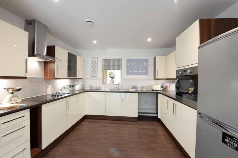 A modern, bright kitchen with a U-shaped layout. The cabinets are a combination of cream and dark wood. Stainless steel appliances, including a cooker hood, oven, refrigerator, and sink, are visible. A white tiled backsplash and wooden flooring complete the space. A framed artwork hangs on the wall above the radiator.