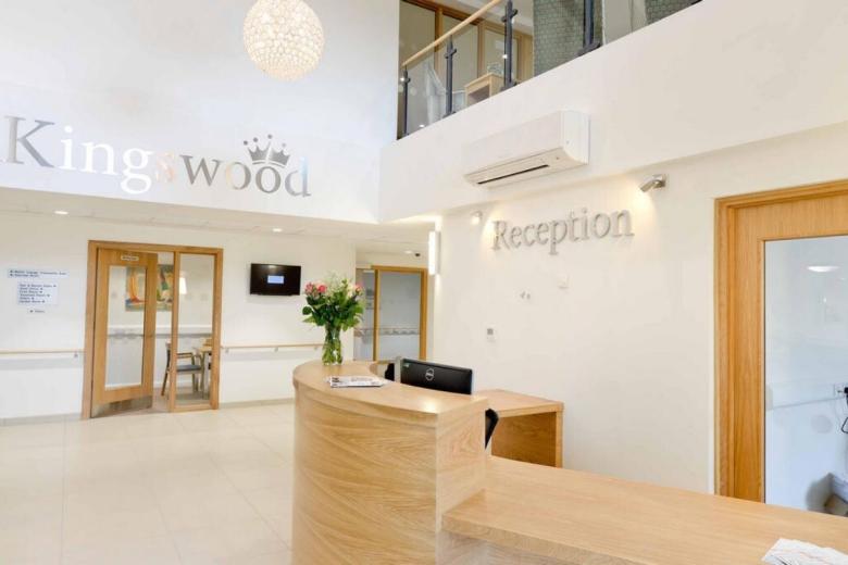 A modern reception area with a light wood reception desk. The desk is adorned with a vase of white flowers. Above the desk, a wall-mounted air conditioning unit is visible. A sign reading "Reception" is mounted on the wall, and the building is identified as "Kingwood." A doorway with artwork is visible in the background, and a television is mounted on the wall to the right.