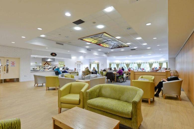 Bright, spacious communal area of a care home with several residents seated in chairs and a small table. A tiled ceiling with a decorative glass panel features above.