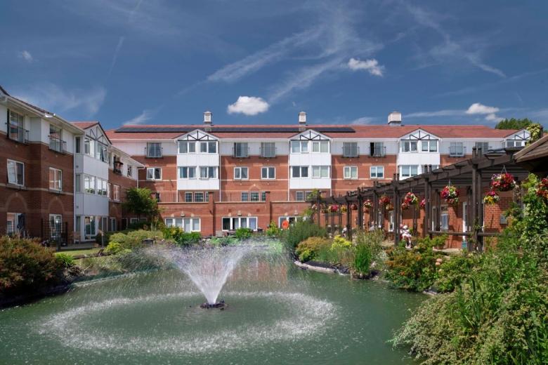 A multi-story brick apartment building with white trim is centered in the image, set within a well-maintained garden. A small pond with a fountain is located in the foreground, surrounded by lush greenery and flowering plants. The sky is blue with scattered white clouds. A covered pergola with hanging baskets is visible to the right of the building.