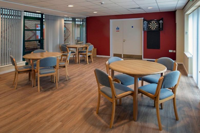 A brightly lit, rectangular room features a wooden-topped circular table surrounded by six matching chairs. The walls are painted a deep burgundy red. To the right, a double door with a white sign is visible, and a dartboard is mounted on the wall adjacent to the door. A vertical blind is positioned in the background window.