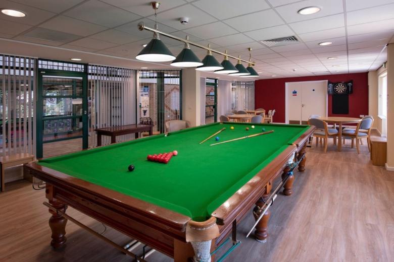 A well-maintained green billiard table dominates the scene, positioned within a spacious room with a light wood floor. The table is adorned with red billiard balls and blue cues. In the background, a table with several chairs is visible, along with a dartboard mounted on a maroon wall. Overhead, pendant lights hang from the ceiling, illuminating the area. The room appears to be a recreational space, likely a common area or games room.