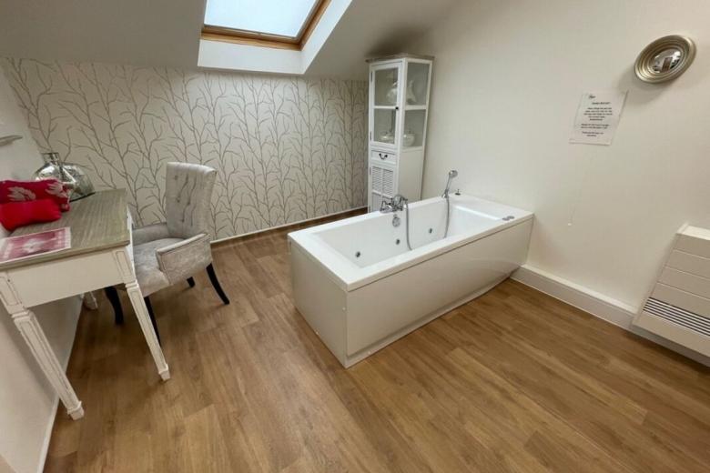 A modern bathroom with a white jacuzzi tub, positioned beneath a skylight. The walls are covered in a grey wallpaper featuring a delicate tree pattern. A small white table with a chair and a glass-fronted cabinet are also present in the room. The floor is covered in light brown wood-effect tiles.