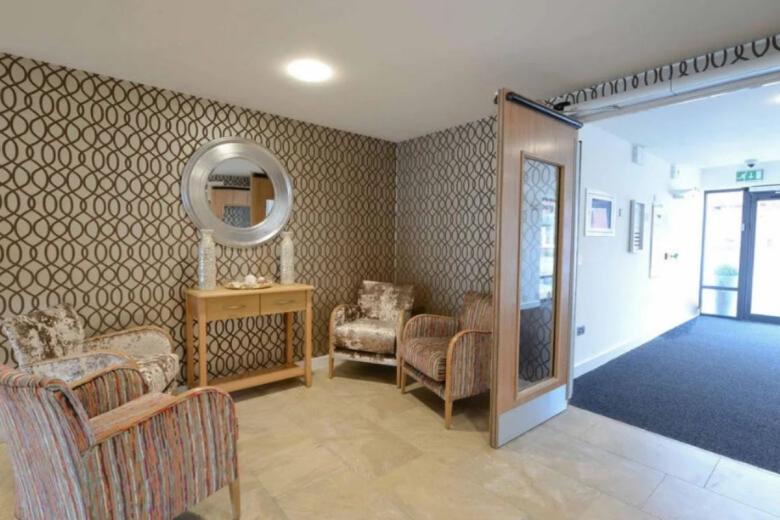 A small waiting area with two patterned armchairs positioned against a wall covered in a repeating geometric pattern of brown, grey, and cream. A small wooden console table with a decorative mirror sits to the left. A partial view of a hallway and a doorway with a green sign is visible in the background.