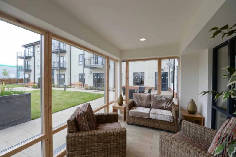 A bright, modern living area features a wicker armchair and a plush sofa positioned in front of a large window overlooking a well-maintained courtyard. The courtyard includes a lawn, trees, and several buildings with balconies. The window frame is made of light-colored wood, and a potted plant sits on a side table. The overall impression is one of a comfortable and inviting space with a pleasant outdoor view.