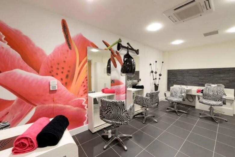 A modern hair salon interior featuring three styling stations. Each station is equipped with a barber's chair and a white grooming table. The walls are predominantly white with a large, vibrant mural depicting a stylized pink flower. One wall is clad in dark grey stone. A black hairdryer is suspended from the ceiling above one of the stations. The floor is tiled in grey.