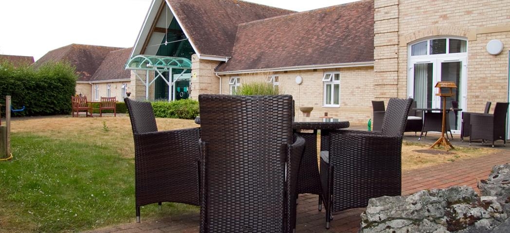 Two outdoor charcoal wicker dining sets are placed amongst the bricked walkways surrounding the grassed area. 
