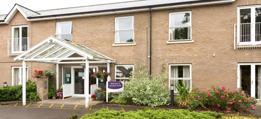Two storey sandy brick-built retirement community located in Cambridgeshire. The entranceway has no step access. The building is decorated by a mixture of mature shrubbery and plants.