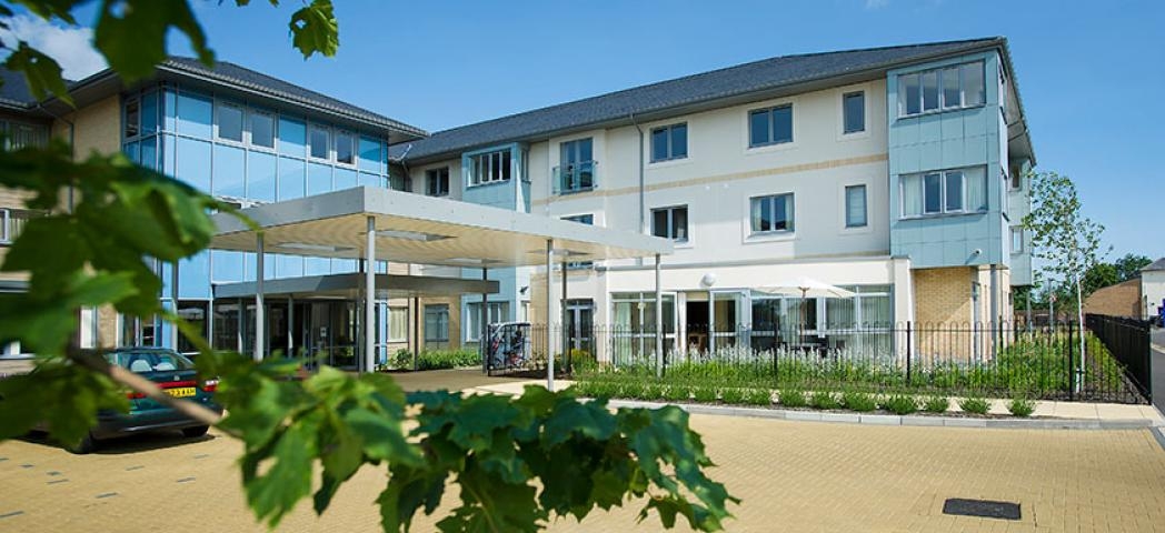 Contemporary, bright retirement living building. The bold blue pops of colour on a selection of the elevations gives a softer approach to the building against the white and grey. A small carpark features at the front of the property. 