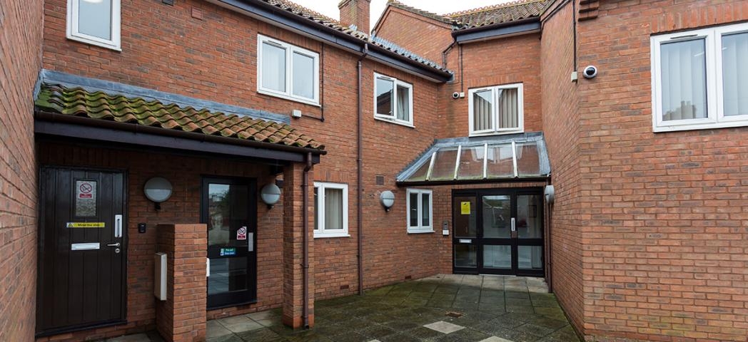 Multi-entranced brick exterior with terracotta clay roof tiles. Multiple security cameras and lighting. Accessed by a clean concrete tiled walkway to Drawbridge House.