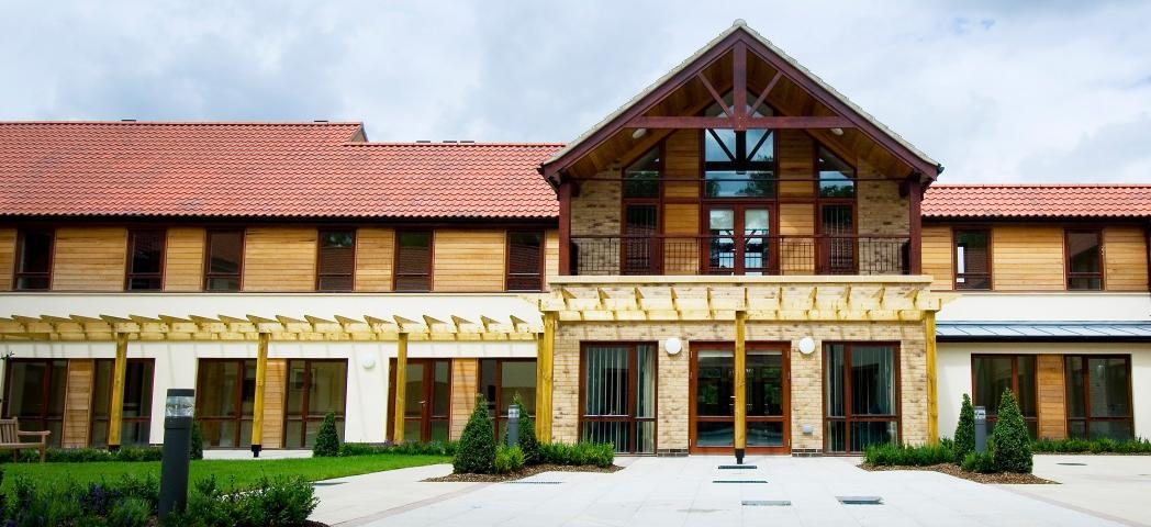 A luxurious property with a grand entrance and first storey balcony overlooking the white slabbed walkway. The property has been built using a mixture of natural materials such as brick, wooden panelling, and wooden beams. Small shrubbery plants line the perimeter.