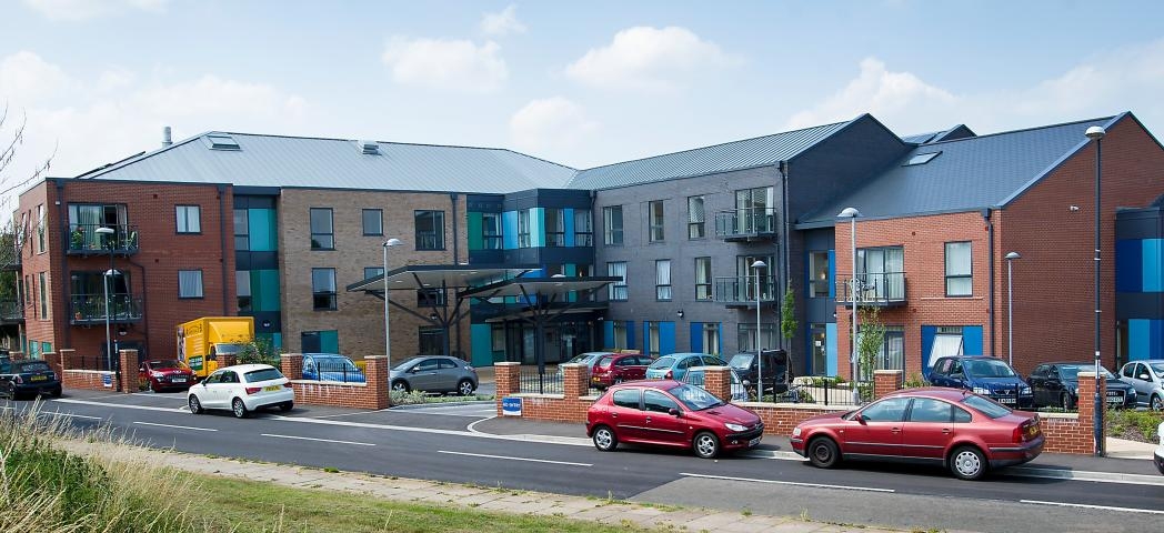 Located on a quiet corner plot the bold retirement living community façade consists of red, stone and grey brick and blue panelling on the larger exterior windows. Parking is located around the perimeter of the building.