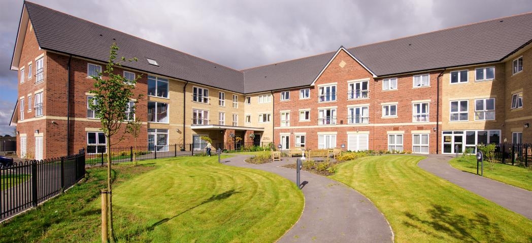 The newly built, yellow, and red bricked, three storey retirement living property set behind winding pathways which lead to a circular outdoor seating area as well the property and lovely grassed areas.