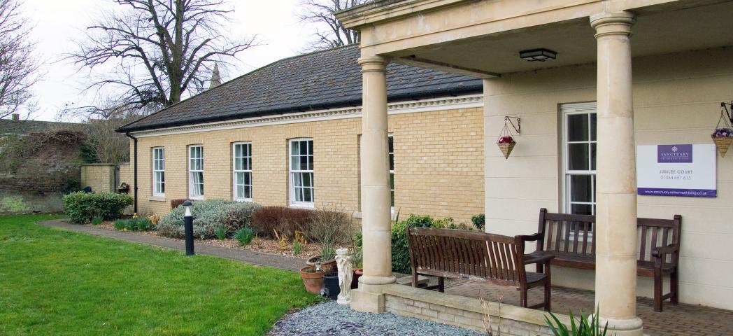 A period designed stone built building is home to the entrance of the property, the entrance is covered by a stone built porch with chunky columns supporting the roof. Two wooden benches sit underneath looking out onto the gardens. The rest of the building is built with soft coloured yellow bricks and long rectangular windows. 