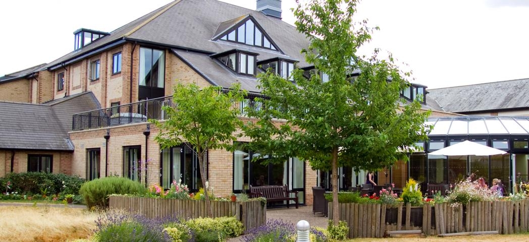An idyllic brick built property with mass amounts of over sized windows and added skylights, set amongst a brick paved patio area and parasol seating enclosed in by wooden logged planters and colourful plants. 