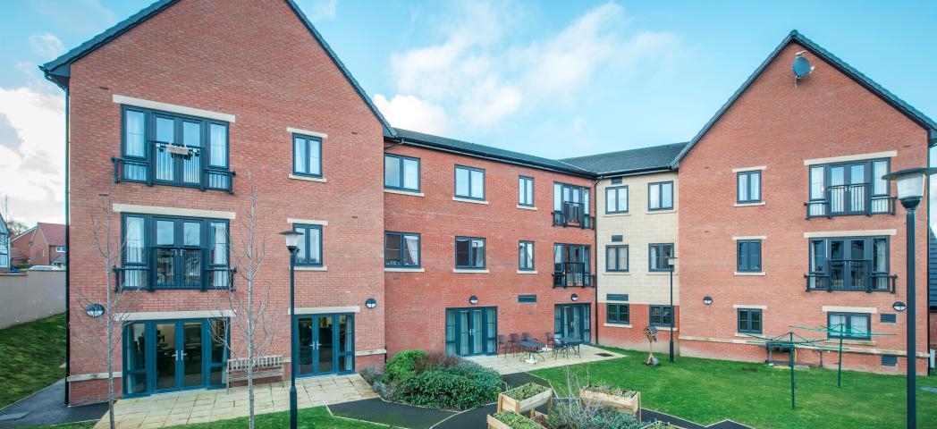 A modern red bricked new build retirement property. The building is built as a three storey corner plot with a small patio area along the left hand side of the property hosts an array of seating options from a wooden bench or metal chairs and circular tables. The patio leads into a circular tarmacked area with wooden planters and shrubbery, the rest of the garden is covered in grass.  