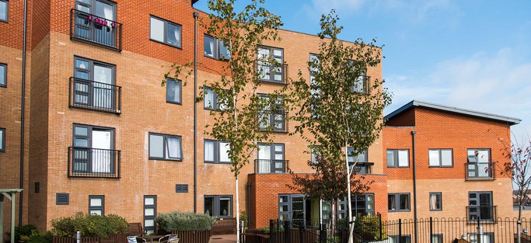 Beautiful contemporary apartment building contrasting dark grey window frames with the multitoned red bricked exterior. The outdoor area offers metal seating as well as numerous wooden benches all looking out to the newly planted trees and garden area.