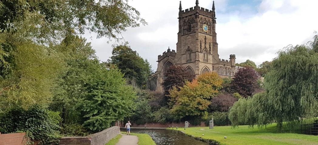 Canal footpath with grass verge and an array of mature trees with a 15th century church towering above.