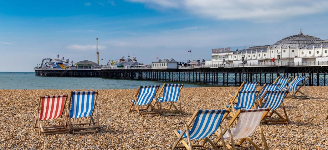 Striped wooden deck chairs placed in pairs on a pebbled beach facing out towards Brighton pier and the sea.