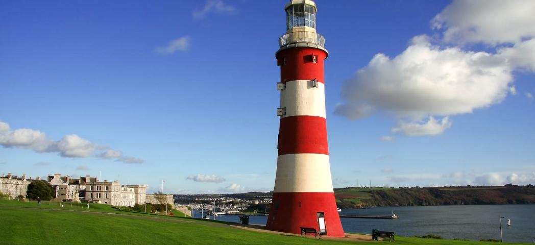 A bold red and white striped lighthouse placed on a grassy cliff, overlooking the Plymouth sound and city.