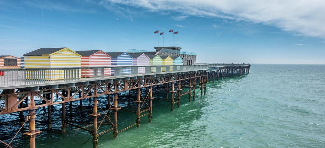 Multiple vibrant pastel and white striped beach huts lining either side of the Hastings pier, contrasting with the teal sea.
