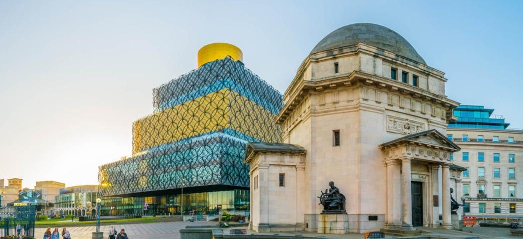 Stone built memorial hall with a small well kept and vibrant garden central to a large pedestrian walkway. In the background a four storey rectangular building with geometric pattern on the outside, alternating between a gold and blue glass colour scheme.
