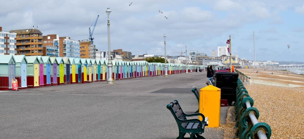 Concrete promenade walkway, to the left lined with a bold multi-coloured petite beach huts and to the right a shingled beach with multiple benches for a sea view.