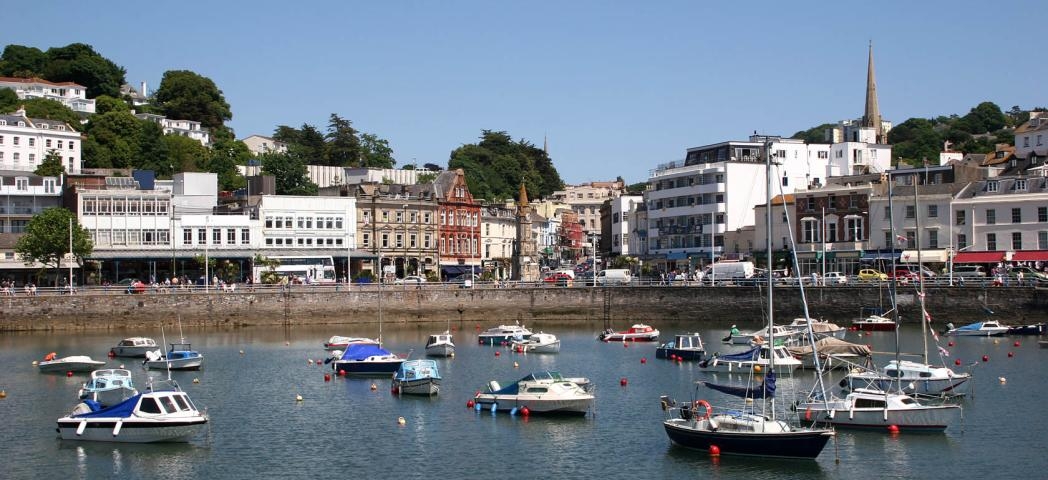 A busy harbour with numerous smaller boats decoratively displaying colourful buoys. A mix of properties cascade down from the hills towards the harbour.