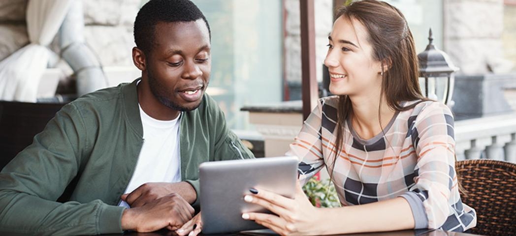 A male being shown something on a tablet style device by a woman, whilst sat next to each other at a table