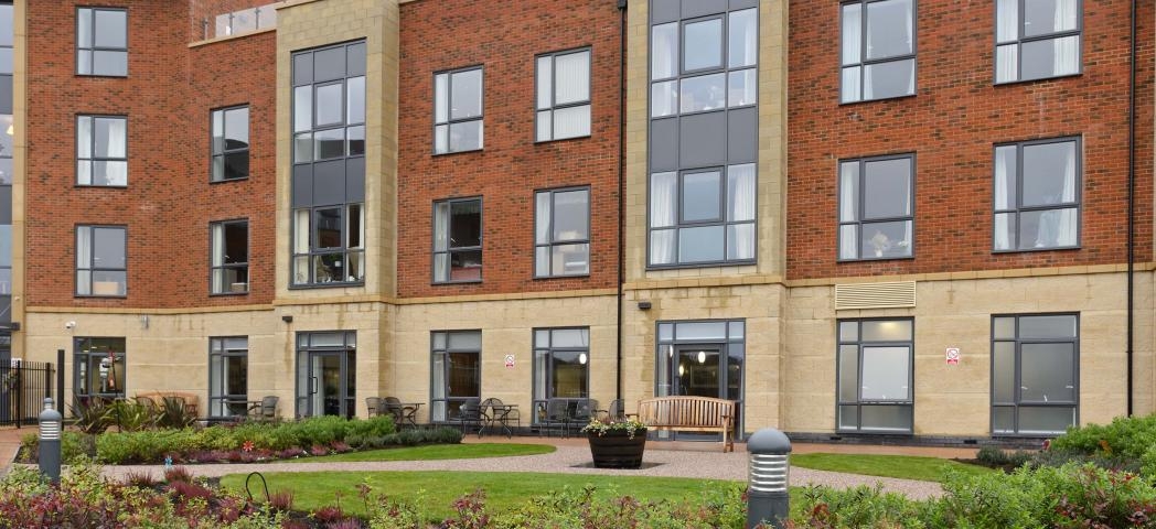 The four storey retirement community. The base of the property is built all in yellow brick, the further three storeys are built using a reddish brown brick with the protruding properties also built with the yellow bricks producing vertical stripes up the property. A outdoor rooftop balcony is seen on the right hand side of the property.