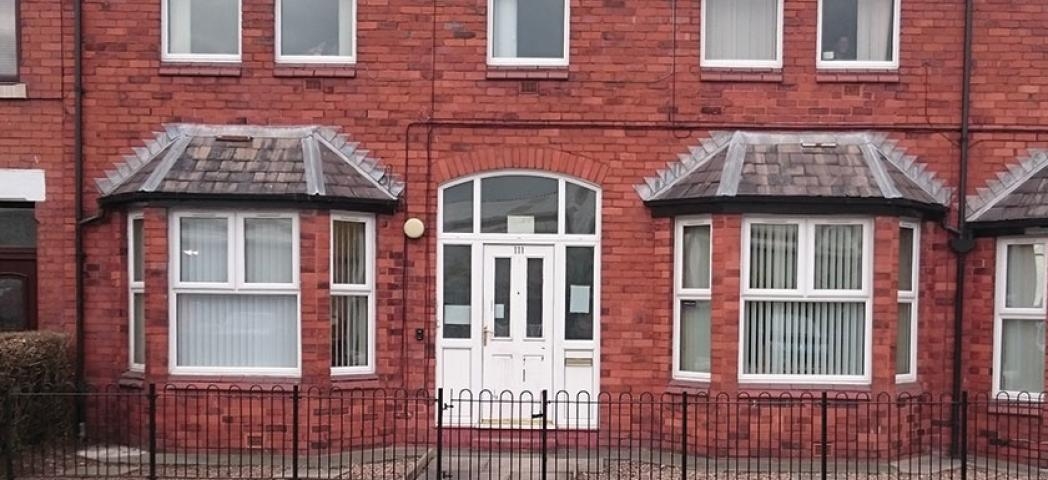 A red brick two storey building with bay windows on the ground floor set on a main road with a low black metal fence and gate in front of the building