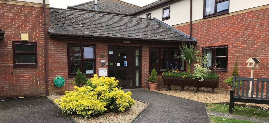 Multi elevational supported living property, built from red brick and a white rendered top elevation. A secure wooden leads into the property surrounded by mature shrubbery and large wooden planters