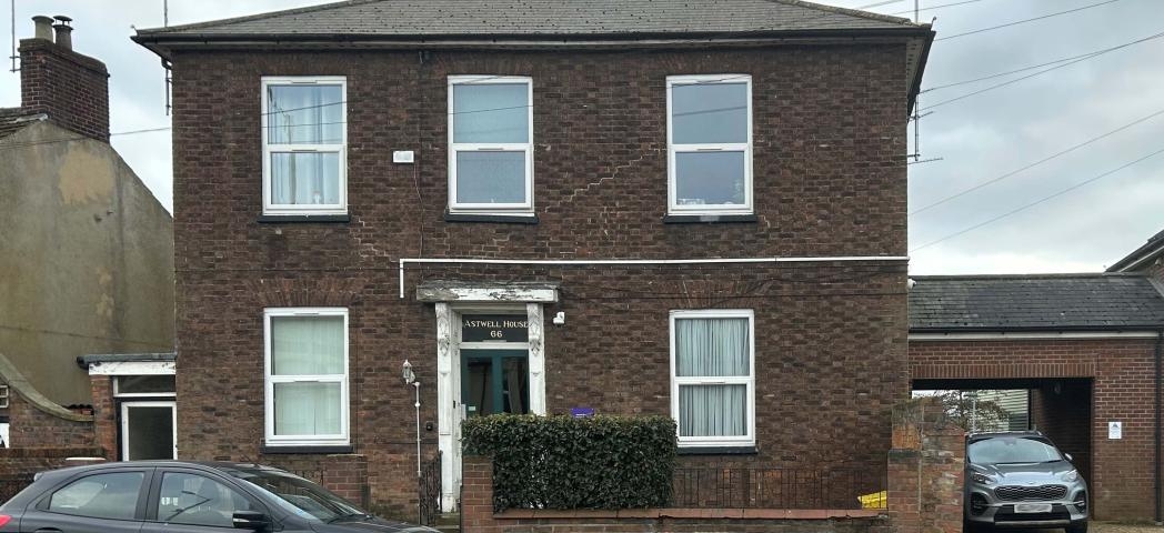 A detached, two storey brown brick built property, with a small brick built wall around the perimeter of the main building. Joined onto the building is a covered opening housing a parking space and access to the back of the property. 