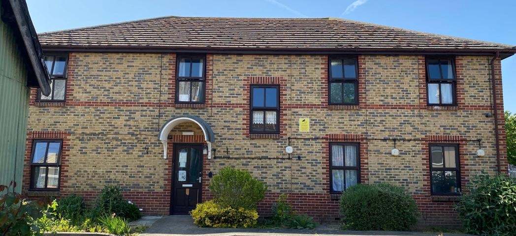 Traditional, elongated, two storey brown bricked property with decorative red brick design around the windows and entrance door. Small, well maintained shrubbery lines the perimeter of the property.