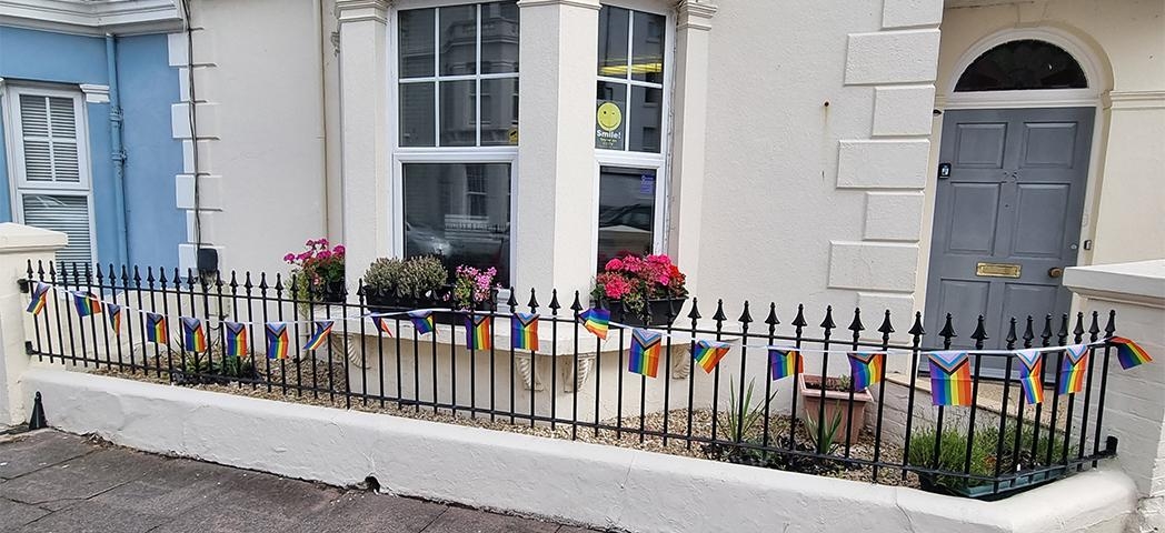 A close up image of the exterior of the building with a grey front door, bay window with sash windows and a black picket style fence out of the front with rainbow pride flags