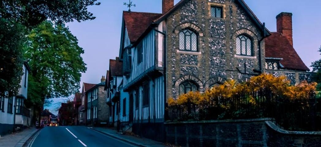 Stock image of a road with a lorry in the distance with a black and white tudor-style building next to a stone wall with metal railings and sunflowers poking over the top