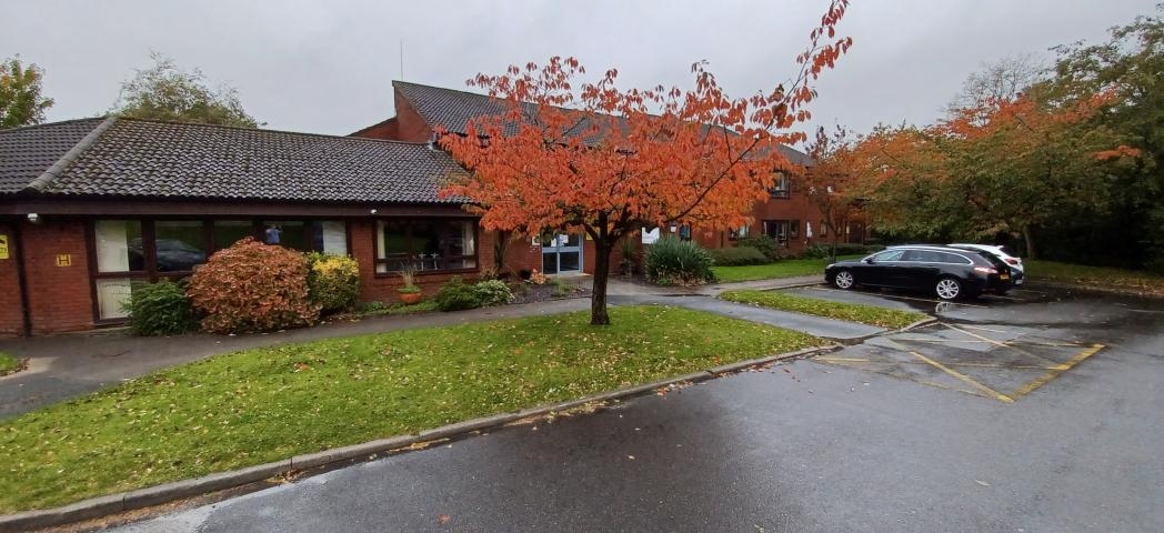 Exterior of red-brick building with a black tiled roof with grass, trees and parking spaces in front