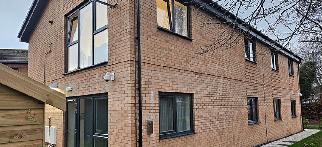 Exterior view of a two-storey supported housing building with light brown brick walls, large black-framed windows, and a paved pathway surrounded by greenery.