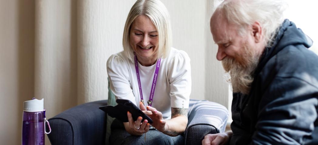 Two people sitting on a couch and laughing, one showing something on a tablet to the other. A purple water bottle is placed on the armrest of the couch.