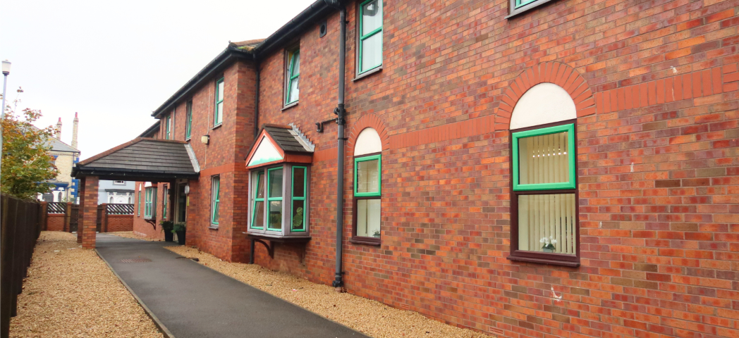 A brick building with green window frames and a sloped roof. The area is surrounded by gravel and a wooden fence, with a cloudy sky overhead.