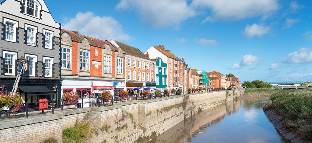 A row of colorful historic buildings along a riverside promenade on a bright sunny day, with reflections in the water